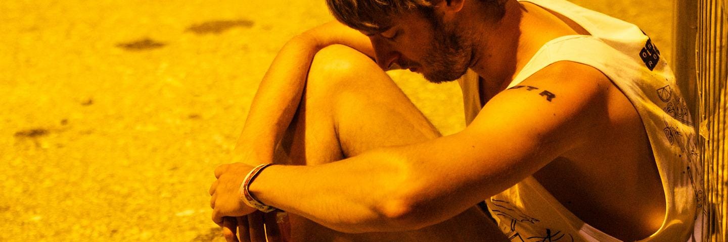 All in shades of yellow, a person sits on the sand with his back against a wall, head bowed, and his arms around his knees. He is wearing brown flip flops, black shorts, and a loose white tank top.