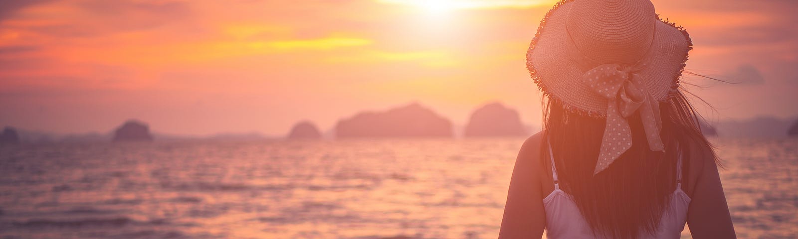 Young woman in a hat, looking at a pink and orange sunset at the beach.