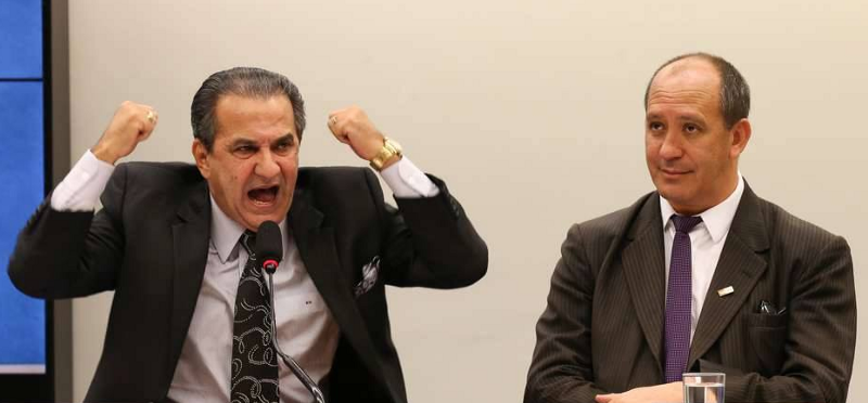 Two politicians participating in a panel discussion; one screams with fists raised in anger while the other looks bewildered