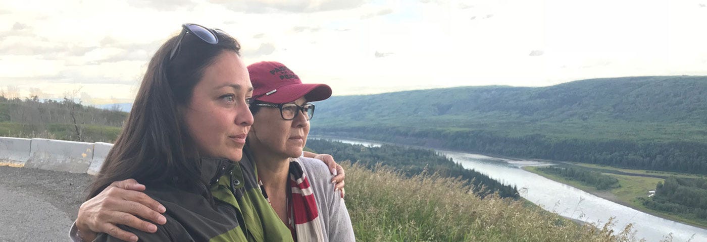 Two women, Heather Hatch and Diane Abel, stand embracing shoulder-to-shoulder while looking over the Peace River Valley.