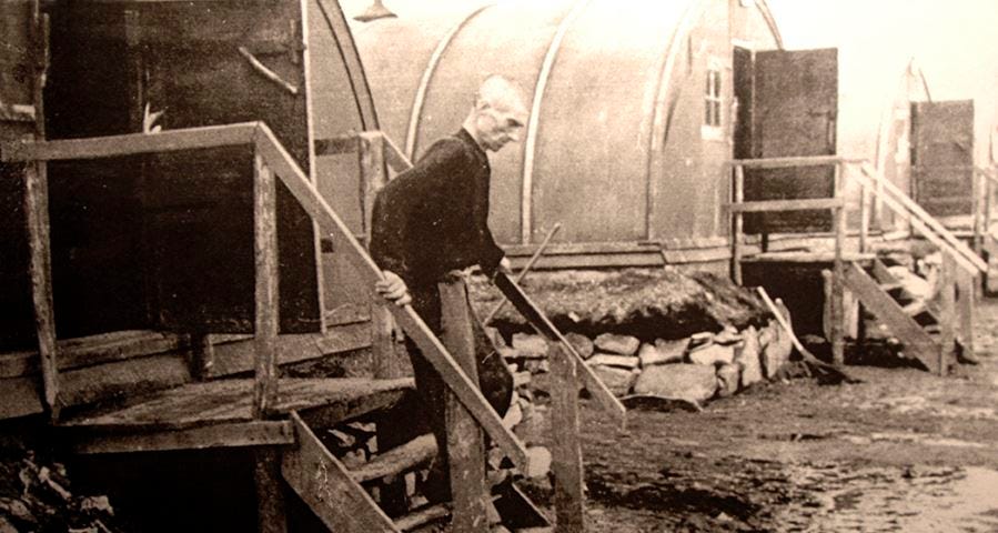 Black white photo of an emaciated looking man carefully walking down steps from a wooden cabin