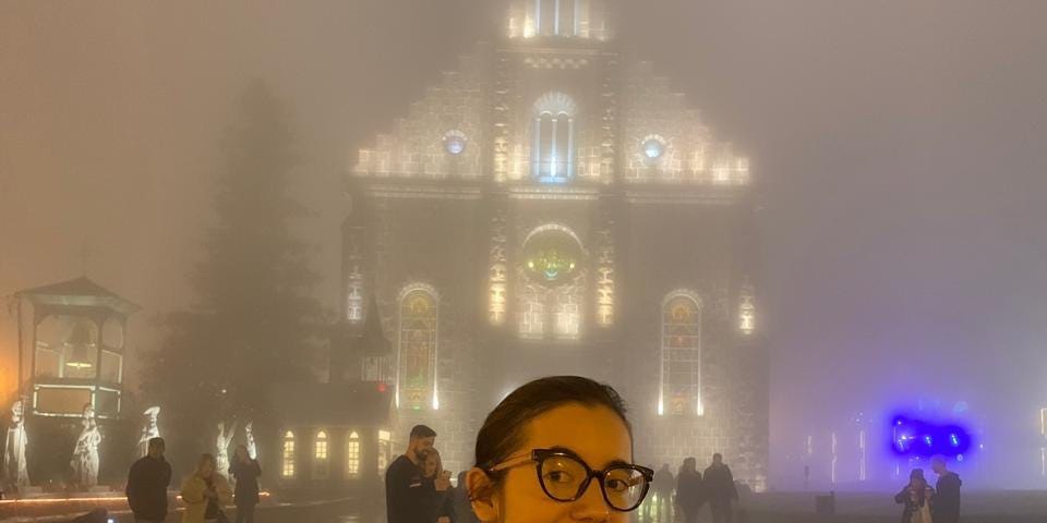 Primeiro plano: eu de cabelo preso, blazer preto sobre suéter preto e óculos. Fundo: igreja coberta por muita névoa.