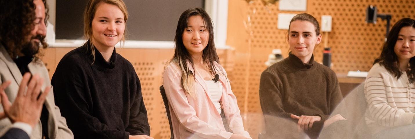 Joe Diaz, Rachael Harkavy, Joyce Yuan, Lancelot Blanchard, and Grace Song are all seated in a row in front of an audience, smiling and listening intently to someone out of the frame.