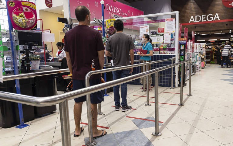 Foto da fila de um supermercado, com marcas de distanciamento social no chão em vermelho e pessoas afastadas 2m das outras