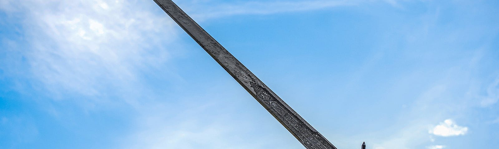 A man’s hand holds an engraved sword into the air. In the backgroudn is a bright blue sky with a few white clouds.