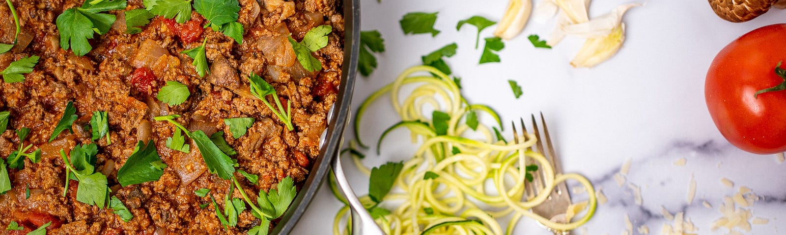 A large pan of cooked ground beef surrounded by garlic, zucchini, tomatoes, and parmesan cheese.