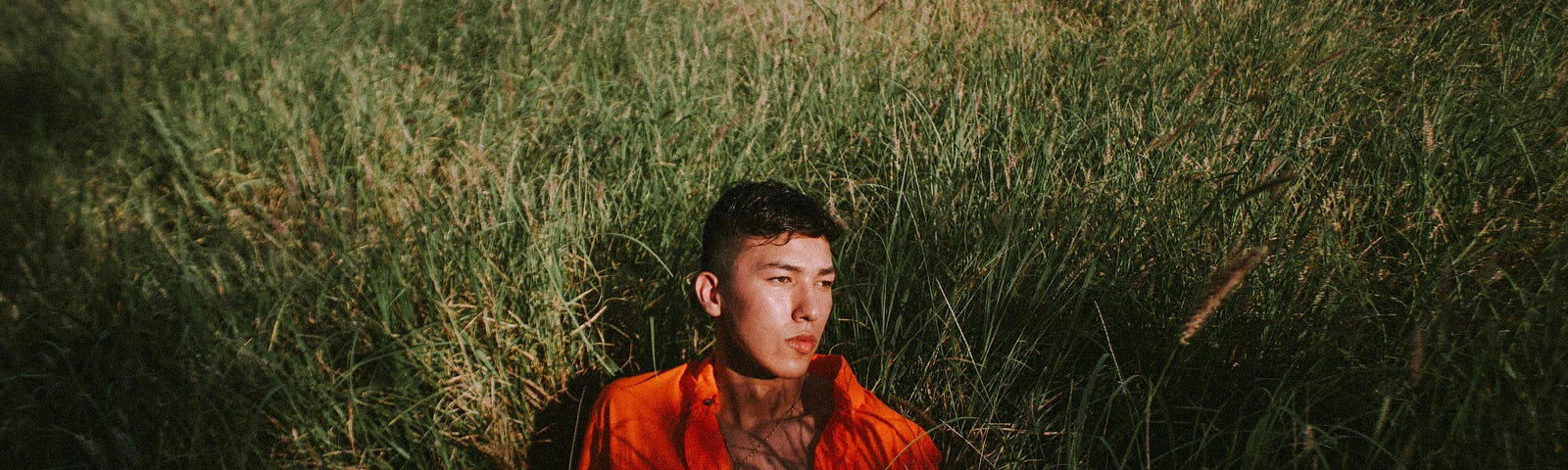 A young man in a field of tall grass, half sitting up and looking off to the side.