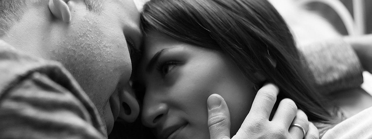 Black-and-white image of a dark-haired woman and half-bald man looking into each other’s eyes, his thumb on her cheek, hand resting in her neck, their foreheads pressed together.