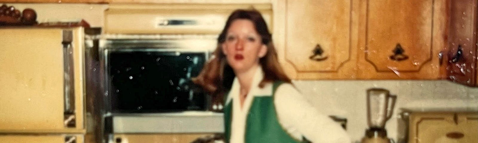 Woman wearing a long green skirt, and matching green vest with a white blouse underneath. She’s in a seventies kitchen with harvest gold appliances and wood cupboards.