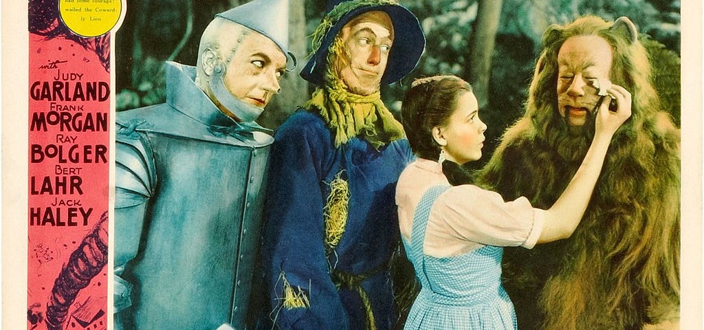 Lobby card from The Wizard of Oz movie, showing Jack Haley in a tin man costume, Ray Bolger in a scarecrow costume, Judy Garland as Dorothy, and Bert Lahr in a lion costume