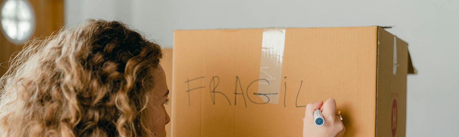 A woman with curly strawberry blonde hair writes “fragile” with a marker on a cardboard moving box.