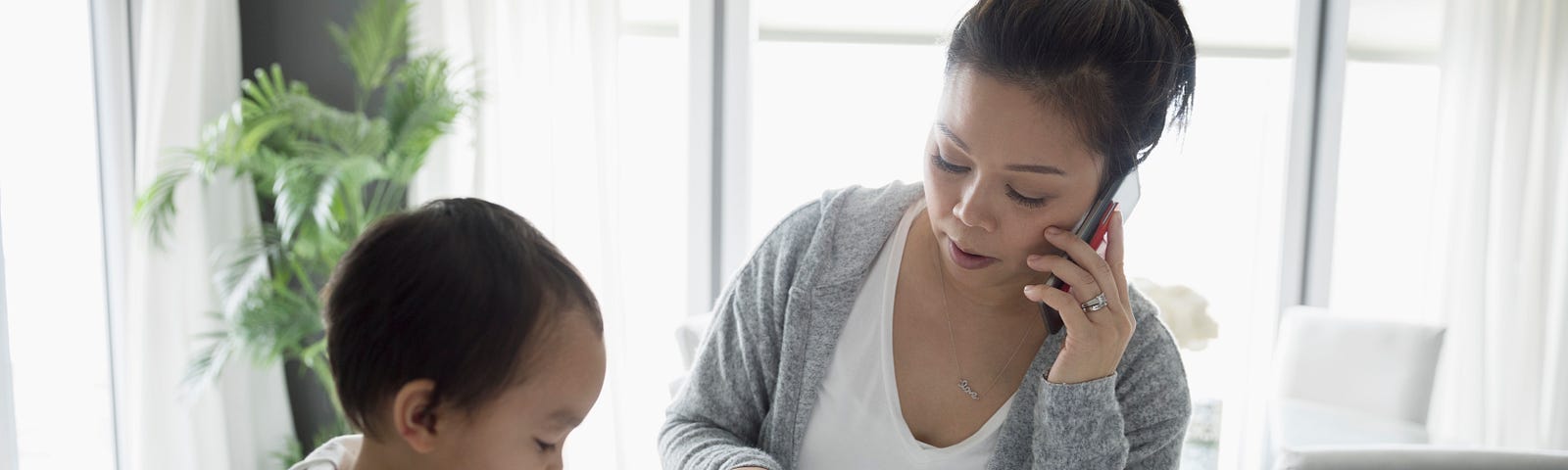 Toddler son and mother paying bills, talking on cell phone in kitchen.