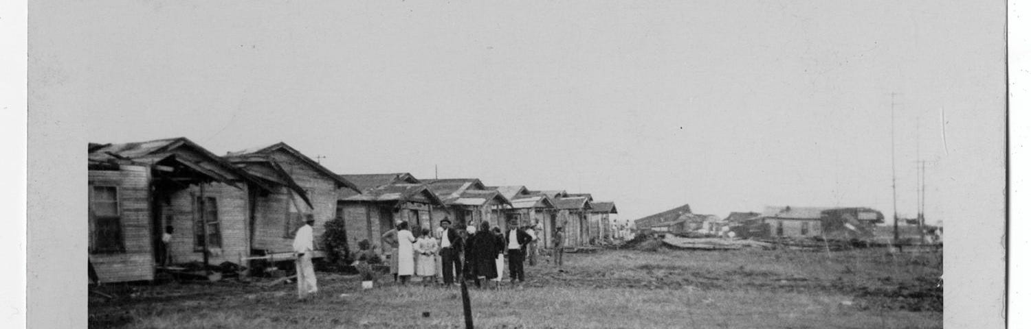 Photograph of a “shantytown” on the outskirts of Port Arthur, Texas. A group of African Americans stand in a group beside a row of dilapidated houses.