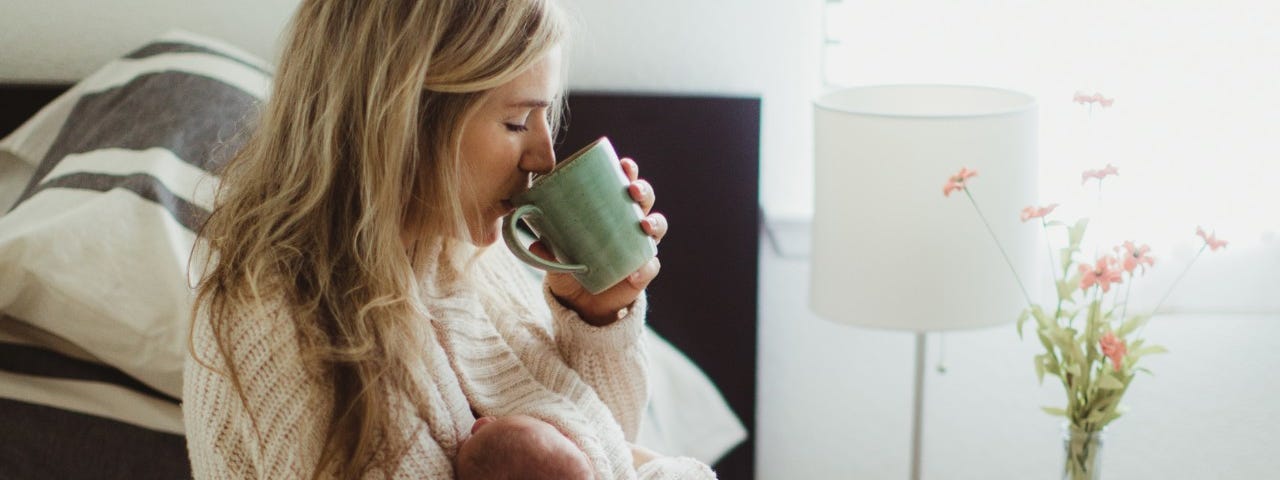 Mid adult woman sitting on bed drinking coffee whilst cradling new born baby daughter