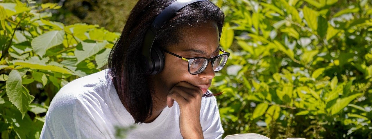 Young woman, wears glasses & a white t-shirt, reads a book. Holds her chin in her hand & appears to be sitting outdoors as the background is leafy