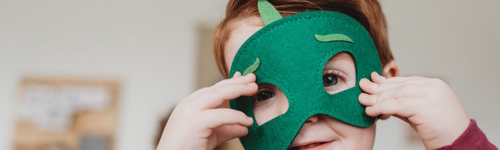 A very small red-haired child in a green cape and dark red top, smiles cheekily from under a green mask.