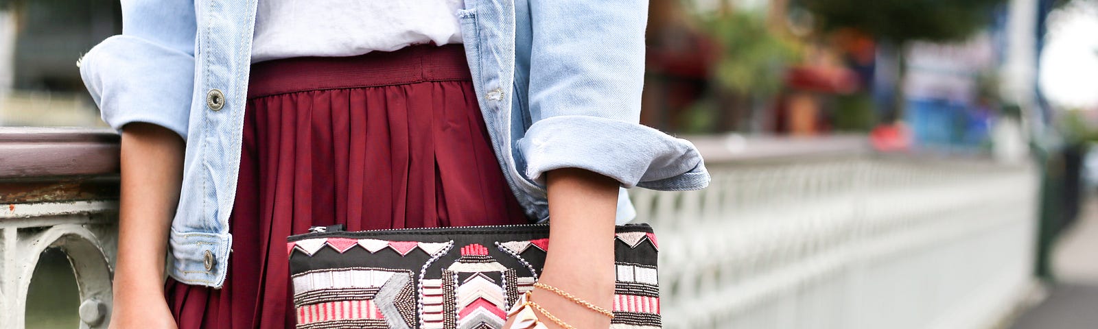 Middle of a person in a short, pleated, pocketless skirt, wearing bracelets and rings, and holding a strapless clutch purse.