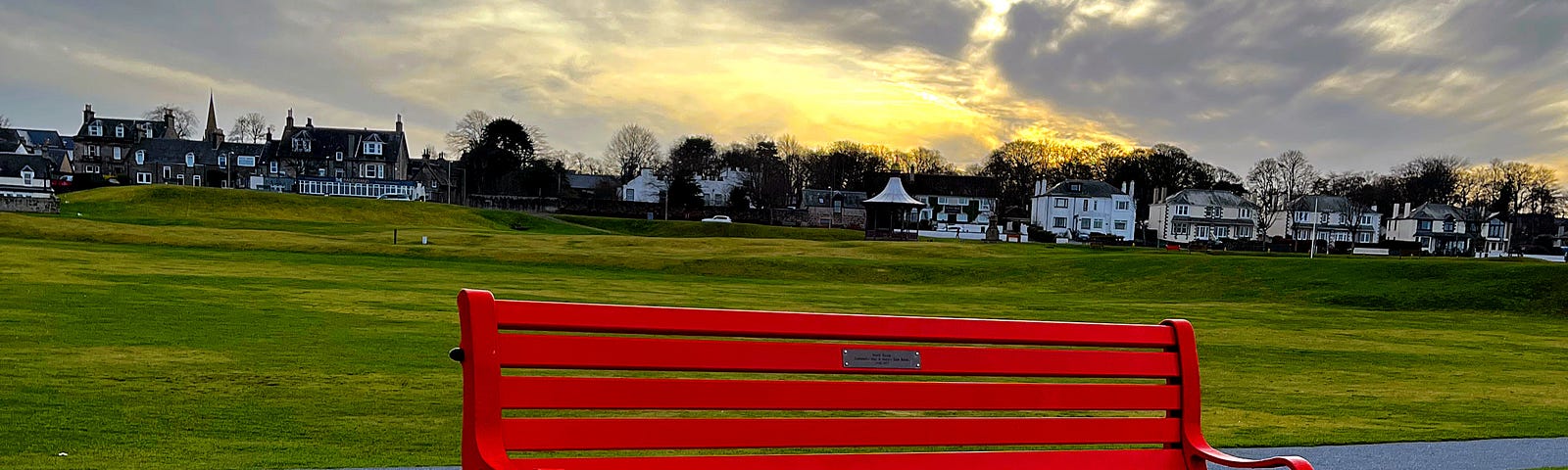 A bright red park bench in a green park at sunset, with family homes in the far background.