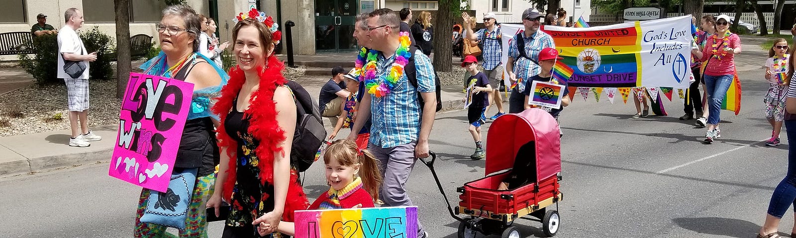 People dressed in bright colours walking in a Pride parade. A girl is holding a rainbow sign that says, “Love conquers hate!” There is a banner for a United Church that says, “God’s Love Includes All.”