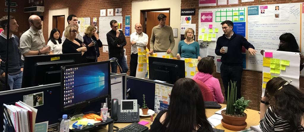 A group of JRF team members stood up in an open plan office presenting a project show and tell to colleagues.