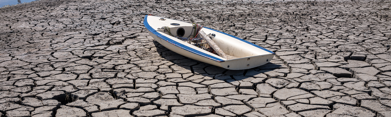 A small white boat stranded on a cracked and dry lakebed, surrounded by parched earth with deep fissures, symbolizing severe drought conditions and water scarcity