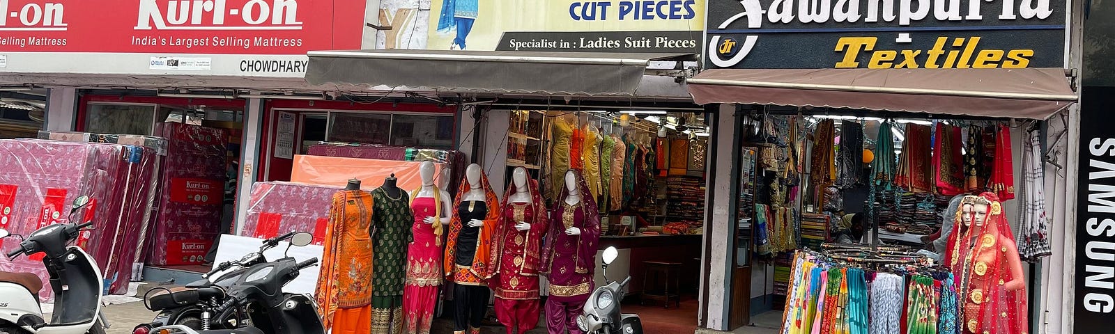 A street in India with clothes shops that display mannequins outside dressed in Indian embroidered clothes. The signboard says, “India Cut Pieces” which means cloth cut into pieces ready for sale and ready to be tailored, usually around 2 meters or three.