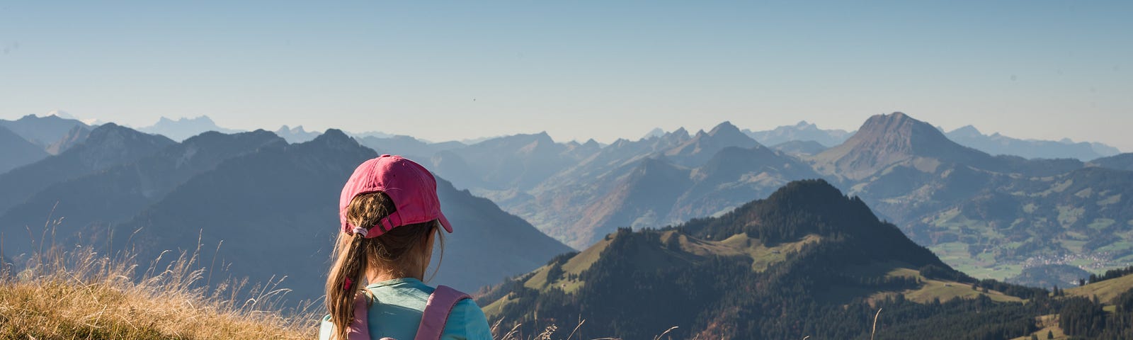 A young girl facing away from the camera and wearing a pink baseball cap and a pink backpack sits on a hill looking out onto a row of mountains that stretch into the distance.