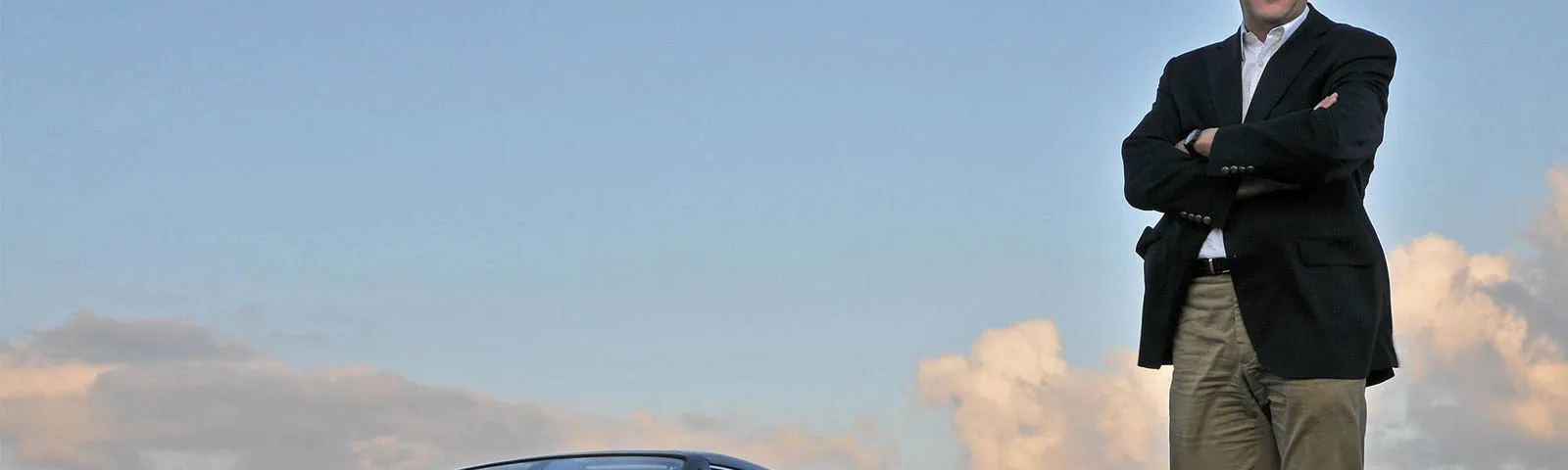 Imagen de Ian Cameron, diseñador de BMW y Rolls-Royce, con los brazos cruzados apoyado en el capó de un automóvil Rolls-Royce. El vehículo está estacionado con un cielo despejado al fondo durante lo que parece ser el atardecer o amanecer, dado el tono del cielo. La placa del vehículo es visible y muestra la inscripción ‘HX08 BHA’.