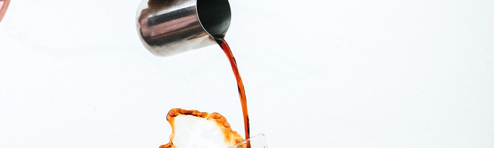 Coffee being poured from a metal jug into a small glass, splashing out over the top and over the hand of the outstretched arm holding the glass.