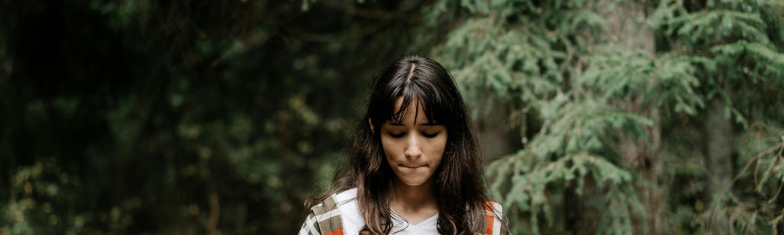 A young brunette teenager wearing a plaid jacket over a white t-shirt looks down at her hands with uncertainty. She’s standing in a forest.