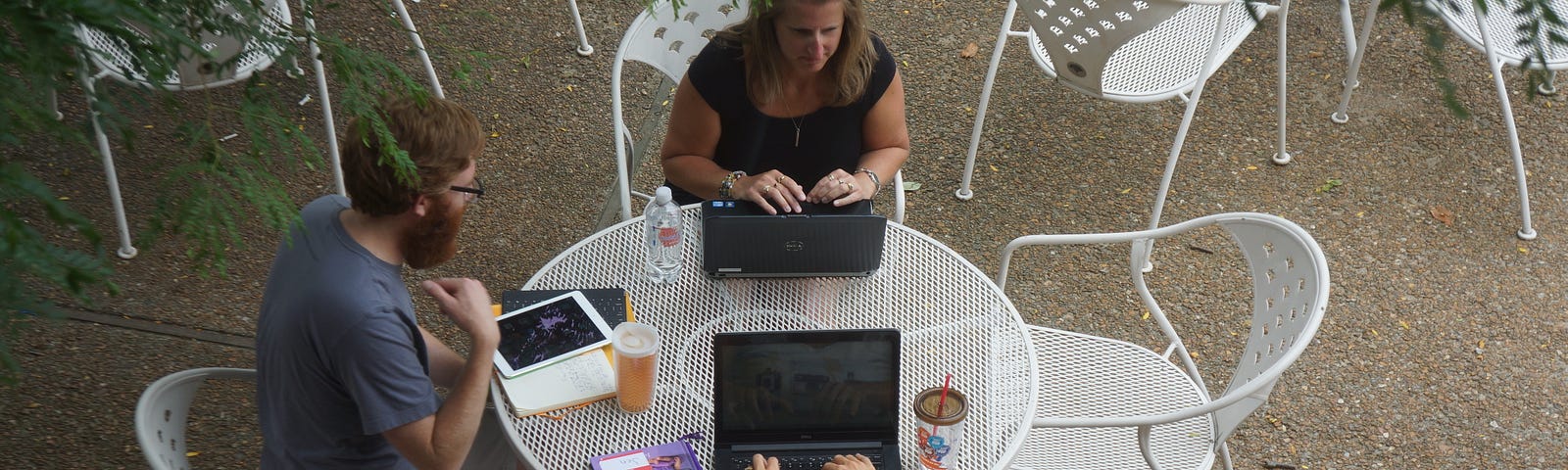 A man and two women sitting around an outdoor table, talking. On the table are books, computers, and writing materials.