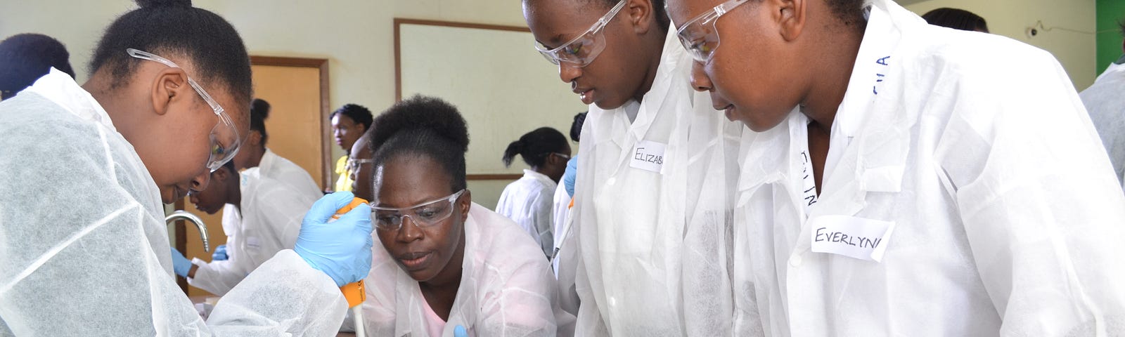 One of four women wearing white lab coats, goggles, and light blue disposable gloves demonstrates a laboratory technique to three of her fellow researchers.