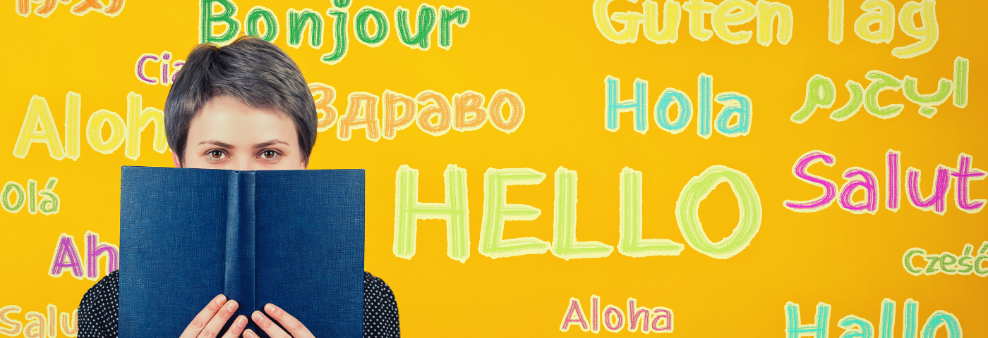 A student woman holding a textbook over a yellow wall written with the word hello translated in different languages.⁠