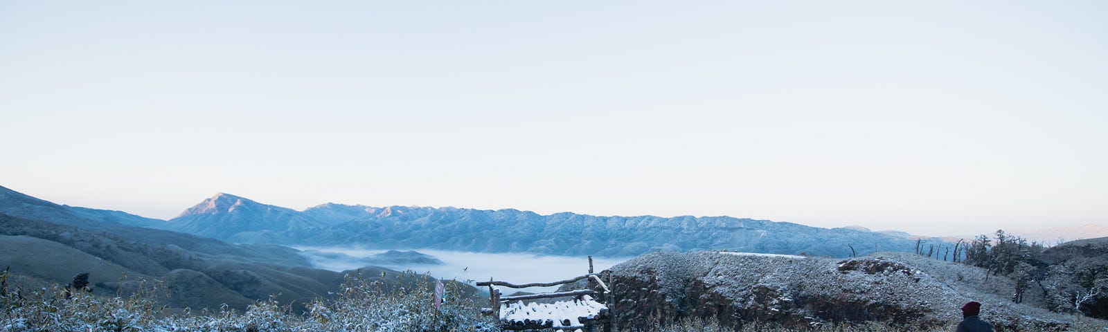 A range of mountains and a lake covered in snow. A small wooden bridge to cross. A person is sitting on a wooden plank.