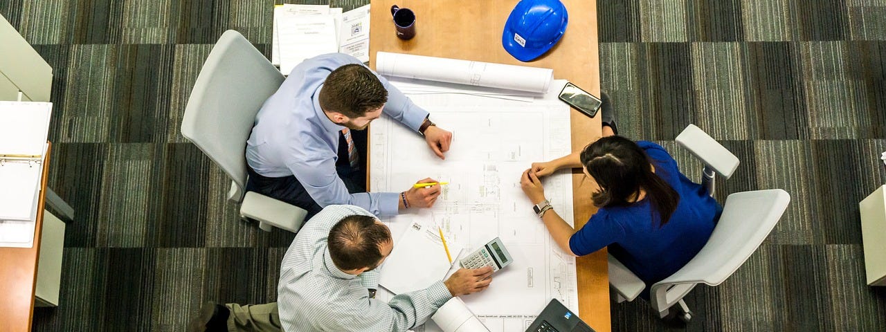 A table with professionally dressed people. Two are on one said on one is on the other. There are drawings, a calculator, laptop, hard hat and other rolls of what are likely to be drawings or plans.