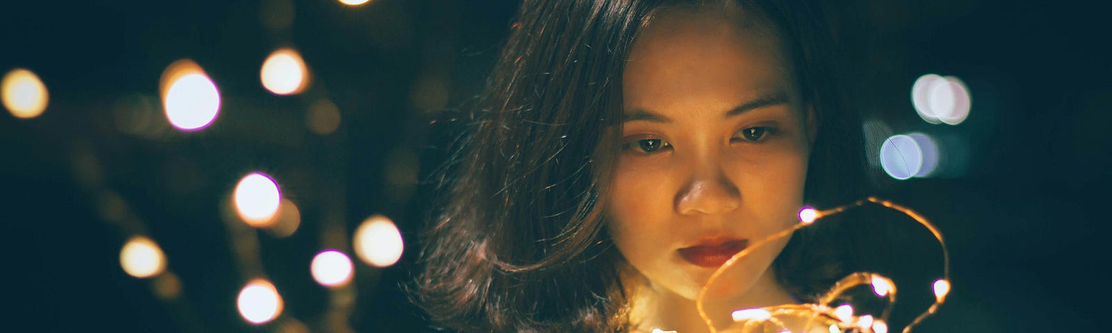 Asian woman gazes at a string of fairy lights in her hand; they cast a soft glow on her face.