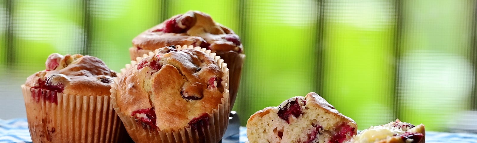 Four delicious sourdough cranberry nut muffins against a spring green background. One muffin in the foreground is cut in half and buttered.