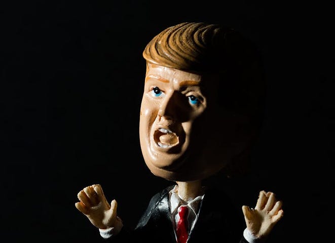A Donald Trump puppet with jazz hands is spotlighted in a black background.