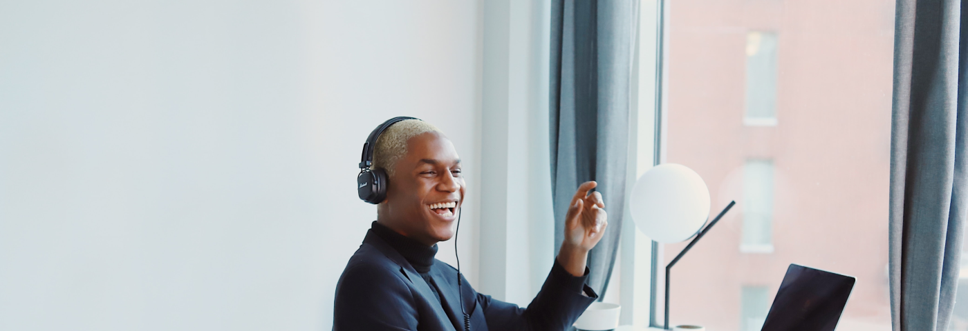 a black man with white hair sitting in front of a laptop, wearing headphones, laughing