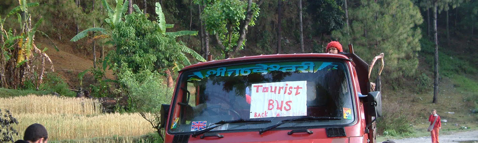Eight men are pushing the front of a red pickup truck with a sign in the windshield that says “Tourist Bus.” There are trees along the side of the road.