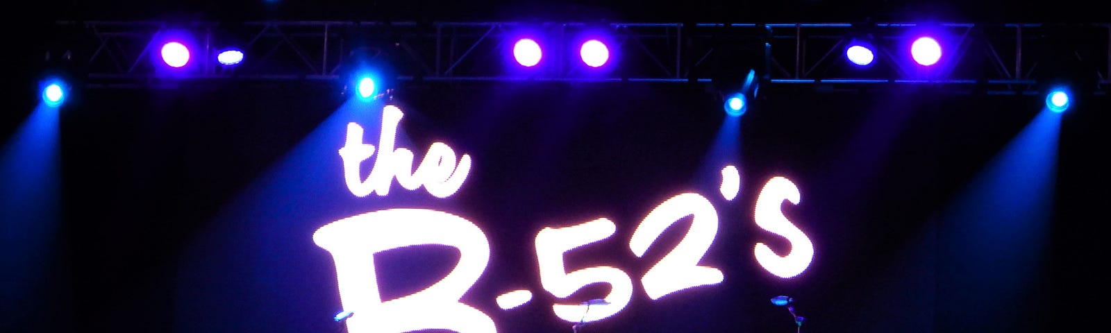 Stage with lit with spotlights and projection of B-52’s logo behind drum kit.