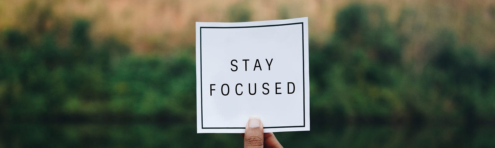 A hand holds a card in front of a lake which reads, “Stay focused.”