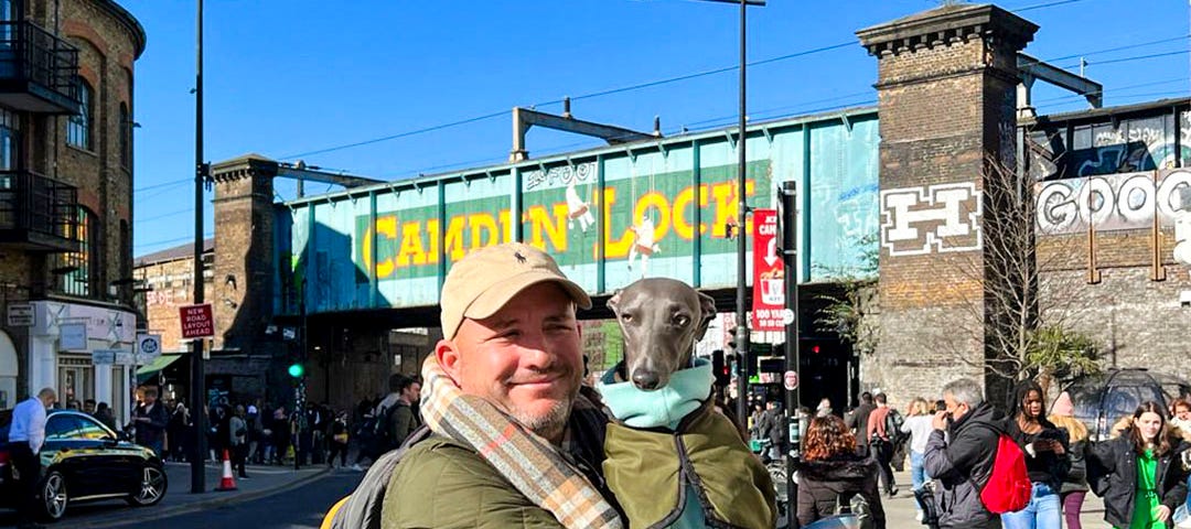 A man in a green jacket is holding a whippet, also in a green jacket, in front of Camden Lock in London