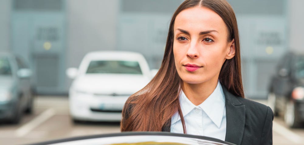 Woman in business suit and long hair parted in middle gets out of her car in parking lot. She holds the door and looks thoughtful, as if trying to remember the parking space she chose.