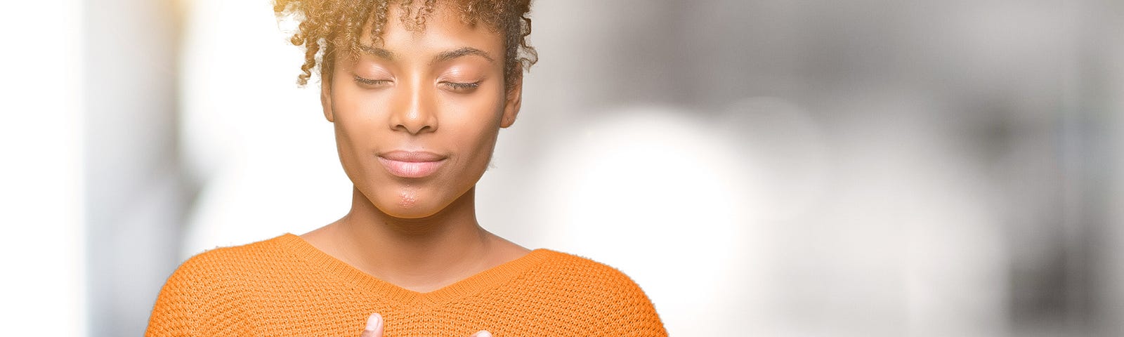 An African American woman stands smiling with her hands on her chest. Her eyes are closed and she has a calm expression on her face.