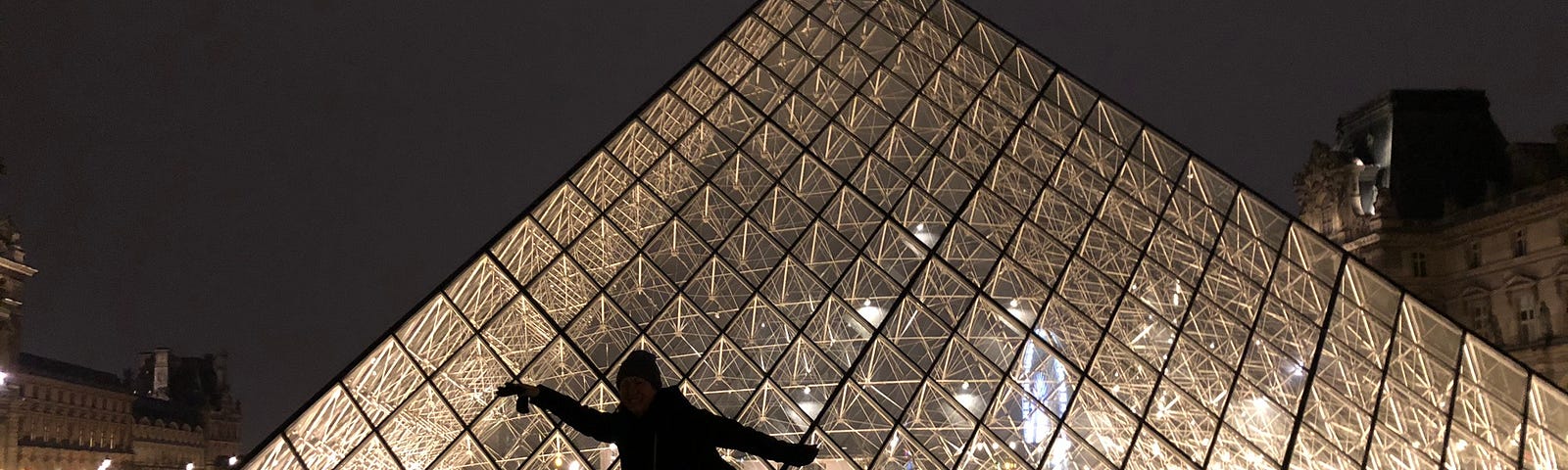 Photo taken at night of the Louvre Pyramid with a woman posing in a silly fashion, backlit by the lighting inside the pyramid.