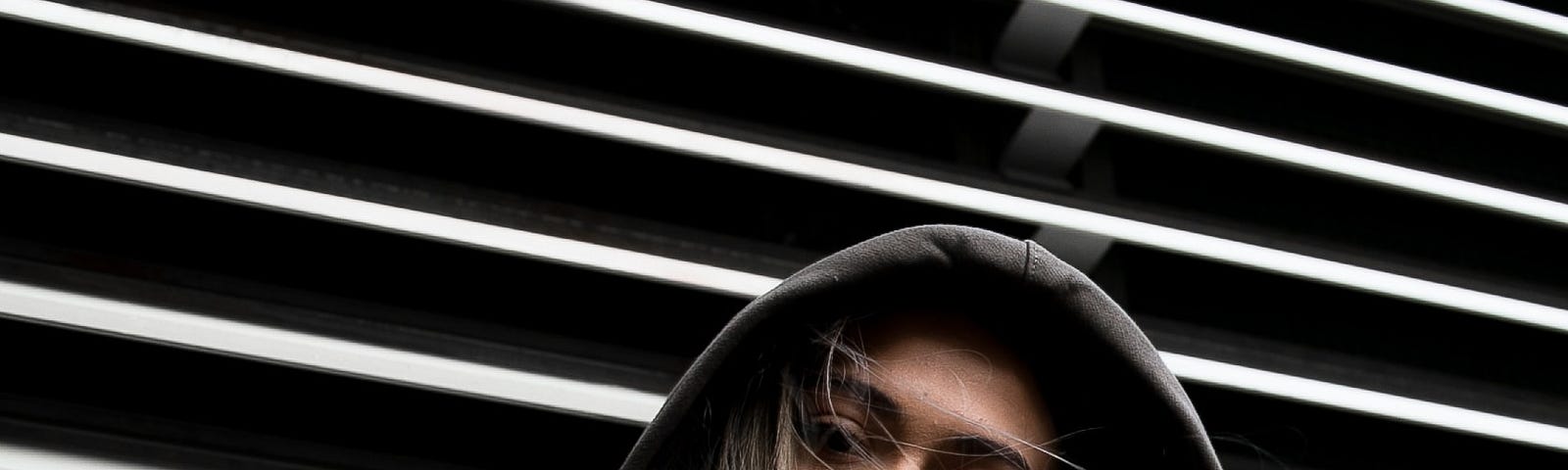 A woman in a hoodie standing in the lower section of Venetian blind window.