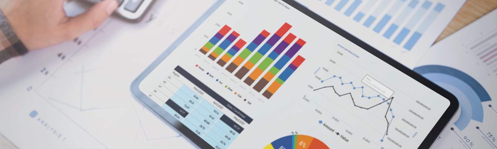 Uma mesa com alguns gráficos impressos sobre ela, onde uma pessoa está estudando um dashboard com diversos tipos de gráficos em um tablet e faz algum cálculo em uma calculadora ao lado.