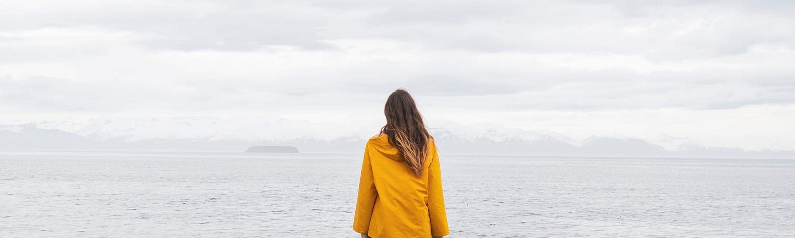 A woman in a yellow coat looking over out into a body of water.