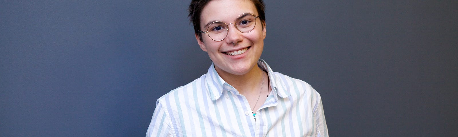 Person with short brown hair and glasses standing in front of a grey background.